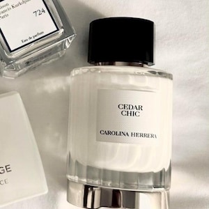 May include: A Carolina Herrera Cedar Chic perfume bottle with a white cylindrical body, a black cap, and a silver base. Next to it is a Maison Francis Kurkdjian 724 eau de parfum bottle with a clear glass square design and a white label.