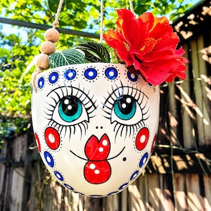 May include: A white ceramic hanging planter shaped like a face with large teal eyes, long black eyelashes, red circular cheeks, and red lips. The planter features blue and black dot patterns along the rim and is suspended by a rope with wooden beads.