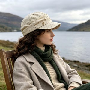Puede incluir: Una gorra de estilo newsboy en color crema con un patrón de pequeños puntos marrones y un detalle de botón metálico en el lateral. La gorra presenta una visera estructurada y un diseño de corona fruncida, con un aspecto clásico y tonos neutros.