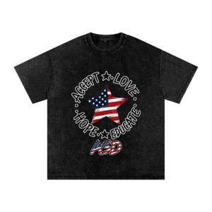 May include: A black mineral-washed short-sleeve t-shirt featuring a central graphic of a star with the American flag pattern. Surrounding the star is the text ACCEPT, LOVE, HOPE, and EDUCATE, with the letters ASD at the bottom in flag colors.