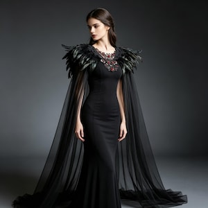 May include: A floor-length black gown featuring a fitted silhouette, a sheer cape with long flowing panels, and a decorative neckline adorned with dark feathers and intricate red and black embellishments that extend over the shoulders.