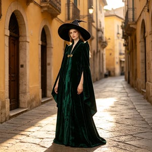 Può includere: Un mantello in velluto verde scuro che arriva fino a terra, caratterizzato da maniche lunghe e ampie e un colletto alto. L'indumento è chiuso sul petto da due bottoni rotondi color oro. Sulla testa è indossato un cappello a punta nero a tesa larga.