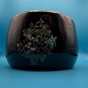May include: A black, rounded bowl featuring a glossy finish and a decorative floral motif. The design includes stylized flowers and leaves in shades of green, blue, pink, and orange, applied with a raised texture against the dark, smooth surface.