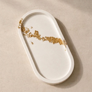 May include: An oval-shaped white tray featuring a smooth, matte finish and a decorative accent of gold leaf flakes scattered across the surface in an irregular, diagonal pattern. The tray has a raised rim and a flat, recessed interior base.
