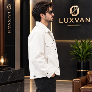 May include: A crisp white denim jacket featuring a classic collar, long sleeves with buttoned cuffs, and a front button closure. The jacket includes two chest pockets with buttoned flaps and is worn over dark trousers, showcasing a clean design.