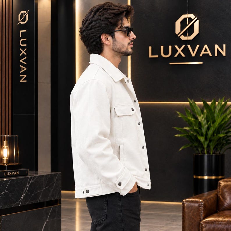 May include: A crisp white denim jacket featuring a classic collar, long sleeves with buttoned cuffs, and a front button closure. The jacket includes two chest pockets with buttoned flaps and is worn over dark trousers, showcasing a clean design.