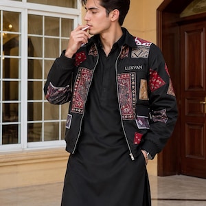 Puede incluir: Una chaqueta negra con cremallera y diseño de parches con paneles de tela estampada en rojo, blanco y marrón en el pecho y las mangas, con el texto LUXVAN en el pecho izquierdo, sobre una túnica negra de manga larga y pantalones negros.