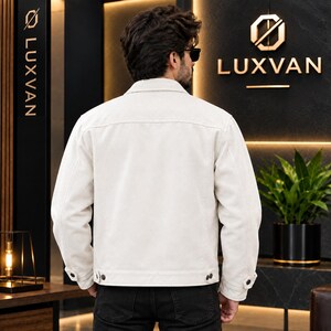 May include: A solid off-white jacket with a classic collar, long sleeves, and buttoned cuffs. The jacket features a straight hemline with adjustable button tabs at the sides. The back view shows a clean, minimalist design with visible seam detailing.