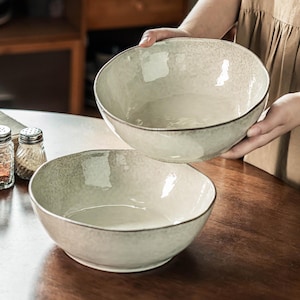 May include: Two round, off-white ceramic bowls with a speckled texture and a dark, uneven rim. The bowls have a slightly irregular, organic shape with a smooth, glossy interior finish and a matte exterior surface, presented as a set of two.