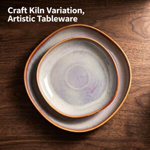 May include: Two round ceramic plates stacked on top of each other, featuring a light purple and cream glaze with a warm brown rim. The text "Craft Kiln Variation, Artistic Tableware" is printed in white at the top of the image.