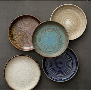 May include: A set of five round ceramic plates arranged in a cluster. The plates feature distinct colors including cream, dark blue, teal, brown, and beige, each with a visible circular grooved texture and speckled finish across the surface.