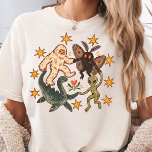 May include: A cream-colored short-sleeve t-shirt featuring a graphic print of a white yeti, a black moth, a green sea monster, and a green alien holding hands in a circle, surrounded by orange stars and a small central floral element.