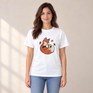 Può includere: Una t-shirt bianca a maniche corte con un'illustrazione grafica di una volpe arancione raggomitolata che legge un libro aperto, circondata da piccole foglie autunnali fluttuanti nei toni del giallo, arancione e verde sul davanti.
