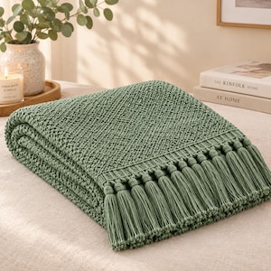 Può includere: Una coperta lavorata a maglia color verde salvia caratterizzata da un motivo intrecciato e testurizzato e da una fila di lunghe nappe abbinate lungo il bordo. La coperta è piegata ordinatamente, mostrando la complessa trama e il colore uniforme.