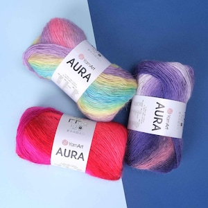 May include: Three skeins of YarnArt Aura yarn with white paper labels. One skein features a pastel rainbow gradient, one is a solid vibrant pink, and one displays a purple and pink gradient. Each label includes brand text and care icons.