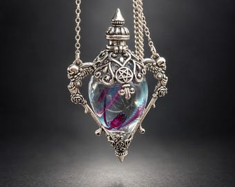 Pode incluir: Um colar com pingente prateado apresentando um frasco de vidro transparente envolto em uma moldura de metal ornamentada. A moldura inclui motivos de caveiras, rosas e um símbolo de pentagrama central. Dentro do frasco, uma pequena criatura roxa e vermelha é visível.