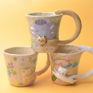 Può includere: Tre tazze in ceramica dalle forme irregolari modellate a mano. Ogni tazza presenta una figura di gatto scolpita sul lato, con la coda del gatto che forma il manico. Le tazze sono decorate con motivi floreali dipinti a mano in colori pastello.