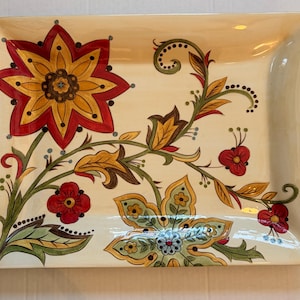 May include: A rectangular cream-colored ceramic platter featuring a large red and yellow star-shaped flower in the upper left corner and a smaller green and yellow floral design in the lower right, connected by green vines and leaves.