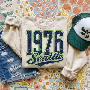 May include: A sand-colored crewneck sweatshirt featuring the dark blue and lime green text 1976 Seattle with a distressed texture. The sweatshirt is paired with blue denim shorts and a white and dark green trucker hat with the text take easy.