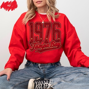 May include: A bright red long-sleeve crewneck sweatshirt featuring the text 1976 and Tampa Bay in a dark red and black athletic font. The design includes small gold star accents between the numbers and a bold, curved underline beneath the text.