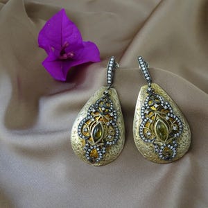 May include: A pair of teardrop-shaped earrings featuring a textured gold-tone finish. Each earring displays an intricate dark metal filigree overlay set with small clear stones and a central olive-green marquise-cut stone. The ear wires are also accented with a row of clear stones.