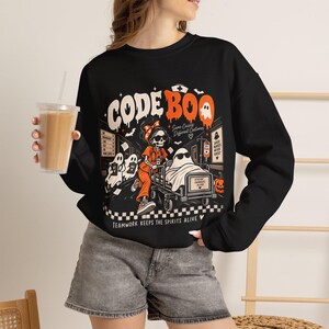 Könnte beinhalten: Ein schwarzes Langarm-Sweatshirt mit einem Grafikdruck, der den Text "CODE BOO" und "TEAMWORK KEEPS THE SPIRITS ALIVE" enthält. Das Motiv zeigt ein Skelett in orangefarbener Kleidung, das eine Trage mit einem Geist schiebt, umgeben von weiteren Geistern.