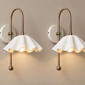 Op de afbeelding: Een paar wandlampen met een witte, gegolfde, klokvormige kap bevestigd aan een gebogen, antiek messingkleurige metalen arm. Het armatuur is aan de muur bevestigd met een ronde witte plaat en is voorzien van een kleine, bolvormige knop.