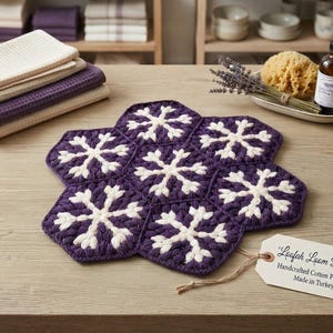 Puede incluir: Un agarrador hexagonal hecho de siete motivos de ganchillo unidos en color morado oscuro con patrones de copos de nieve blancos. Una etiqueta de papel beige atada con cordel dice Loofah Loom Design, Handcrafted Cotton Potholder, Made in Turkey.