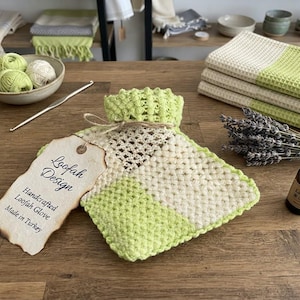 Puede incluir: Un guante de lufa de ganchillo cuadrado con cuadrados texturizados en verde claro y crema, atado con una cuerda a una etiqueta de papel que dice Loofah Design, Handcrafted Loofah Glove, Made in Turkey, junto a una pequeña botella de vidrio marrón.