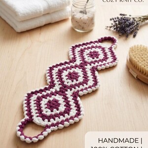 Puede incluir: Un estropajo de espalda de ganchillo largo y rectangular con tres motivos cuadrados interconectados en hilo color ciruela oscuro y blanco. El estropajo tiene un asa circular en cada extremo y el texto COZY KNIT CO. y HANDMADE | 100% COTTON | SPA ESSENTIAL.