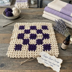 Puede incluir: Un agarrador de crochet cuadrado con un patrón de tablero de ajedrez en hilo color crema y morado oscuro. En la esquina hay una etiqueta de papel beige con el texto Loofah Loam Design, Handcrafted Turkish Loofah Cloth, Made in Turkey, atada con un cordel.