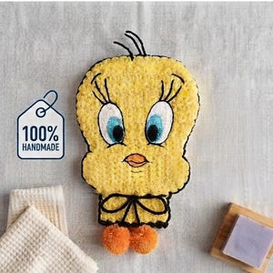 Puede incluir: Un personaje de Tweety tejido a ganchillo en amarillo con grandes ojos azules y blancos, pestañas negras y un pico naranja. Presenta un lazo negro en el cuello y dos pompones naranjas en la base. Se ve una etiqueta blanca con 100% HANDMADE.