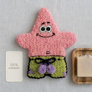 Puede incluir: Una figura de estrella de mar tejida a ganchillo en rosa y verde con grandes ojos blancos, una sonrisa cosida en negro y pantalones cortos verdes con un cordón negro y detalles morados. Una etiqueta blanca dice 100% HANDMADE junto a una bandeja de madera con jabón.