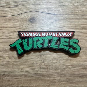 Op de afbeelding: Een Teenage Mutant Ninja Turtles logo-bord met de woorden Teenage Mutant Ninja in witte letters op een rode achtergrond, en het woord Turtles in grote, groene, getextureerde letters met een zwarte omlijning voor een 3D-effect.