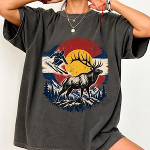 Puede incluir: Una camiseta de manga corta gris carbón con un estampado gráfico de un esquiador y un alce sobre una cresta montañosa. El diseño se sitúa sobre un fondo circular con un sol amarillo y franjas de color rojo y azul.