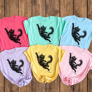 May include: A collection of six short-sleeved crew neck t-shirts in pink, mint green, light blue, lavender, yellow, and light pink. Each shirt features a black ink-style illustration of a fluffy black cat reaching upward with its paws.