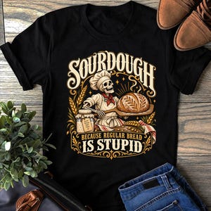 Puede incluir: Una camiseta negra de manga corta con un gráfico de un esqueleto con gorro de chef y delantal, sosteniendo un pan en una pala. El texto dice SOURDOUGH BECAUSE REGULAR BREAD IS STUPID, con un frasco etiquetado Good Things Take Time.