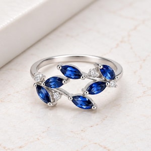 May include: A silver-toned ring featuring a band designed with marquise-cut deep blue stones arranged in a leaf-like pattern, accented with small, clear round stones set between the blue stones along the open-style metal band structure.