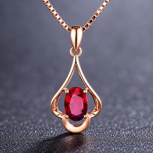 May include: A rose gold pendant necklace featuring a central oval-cut deep red gemstone held by a four-prong setting within a teardrop-shaped frame. The pendant hangs from a delicate box chain, showcasing a polished metallic finish throughout.