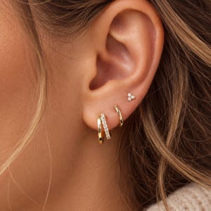 Op de afbeelding: Een set van drie goudkleurige oorbellen gedragen in de oorlel en het kraakbeen. De set bevat een gladde gouden ring, een gouden ring bezet met heldere steentjes en een kleine oorsteker met drie kleine steentjes in een driehoekige vorm.