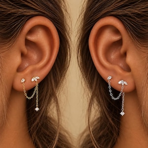 May include: A dual-post earring featuring a cluster of marquise and pear-cut clear stones connected by two delicate chains to a single dangling round stone. The design is shown in both gold and silver metal finishes, each worn in an earlobe.