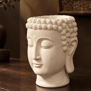 Puede incluir: Una maceta blanca de acabado mate con forma de cabeza de Buda. La escultura presenta cabello rizado estilizado, ojos cerrados, una expresión facial serena y una abertura circular en la parte superior. La superficie tiene una textura similar a la piedra.