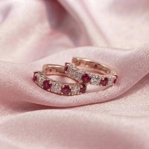May include: A pair of hoop earrings crafted from rose-toned metal, featuring an alternating pattern of round-cut, deep red rubies and clear, sparkling white stones set along the front-facing outer edge of each circular hoop.