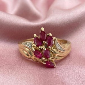 May include: A gold ring featuring a cluster of marquise-cut red rubies arranged in a floral-like pattern. The band is crafted from yellow gold and includes small, clear accent stones set along the shoulders of the ring, adding detail.