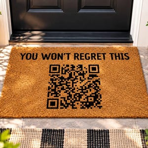 May include: A rectangular tan coir doormat featuring the black text YOU WON'T REGRET THIS printed above a large black QR code. The mat has a coarse, fibrous texture and is positioned horizontally in front of a dark door.