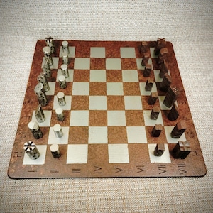 Könnte beinhalten: Ein quadratisches Schachbrett mit einem braun strukturierten Rand, der römische Ziffern von I bis VIII aufweist. Das Brett hat abwechselnd silberne und braune Felder. Eine Seite hat zylindrische silberne Figuren, die andere dunkelbraune geometrische Figuren.