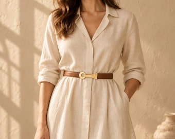 May include: A beige button-down shirt dress with long sleeves and a collared neckline. The dress is cinched at the waist with a thin brown leather belt featuring a gold-toned metal buckle. The fabric has a visible textured weave.