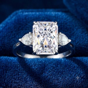 May include: A silver-coloured ring featuring a large, rectangular radiant-cut centre stone held by four prongs. The centre stone is flanked by two smaller, pear-shaped side stones, one on each side, set on a polished, solid metal band.