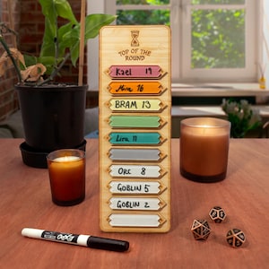 May include: A vertical wooden initiative tracker board with an hourglass icon and the text TOP OF THE ROUND. It features eight horizontal slots with colored inserts labeled Kael 19, Mica 16, BRAM 13, Lira 11, ORC 8, GOBLIN 5, and GOBLIN 2.