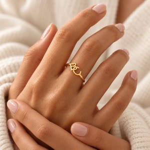 Könnte beinhalten: Ein goldfarbener Ring mit zwei miteinander verbundenen Buchstaben, B und C, die einen zentralen Blickfang auf einem dünnen, polierten Band bilden. Das Design ist schlicht und elegant und zeigt die Buchstaben in einer klaren, serifenlosen Schrift.